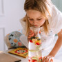 Dziewczynka z karafką wody z owocami ubrana w fartuszek wodoodporny dla dzieci – dzikie kwiaty na błękicie z kołnierzykiem i białą dużą kieszenią. Fartuszek jest wykończony jasnobeżową koronką kostkową. Fartuszek jest wiązany na szyi oraz na pleckach, jak klasyczny kuchenny fartuch.