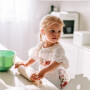 Dziewczynka wałkująca coś na stole ubrana w fartuszek wodoodporny dla dzieci – purpurowy ogród przedstawia piękne, kwitnące polne kwiaty. Białe tło doskonale wydobywa głębię kolorów wzoru. Fartuszek jest wiązany na szyi oraz na pleckach, jak klasyczny kuchenny fartuch. Wykończony jest 1 cm koronką i dużą białą kieszenią z przodu.